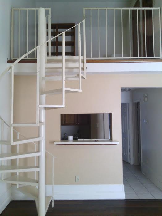 A small room with a white spiral staircase leading to a loft area, a counter window showcasing a cozy kitchen, and light beige walls. This charming property for lease is part of the sought-after Baton Rouge real estate market, ideal for those looking for both comfort and convenience.