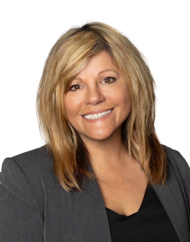 A person with shoulder-length blonde hair smiles while wearing a dark blazer and black top, set against a plain white background.