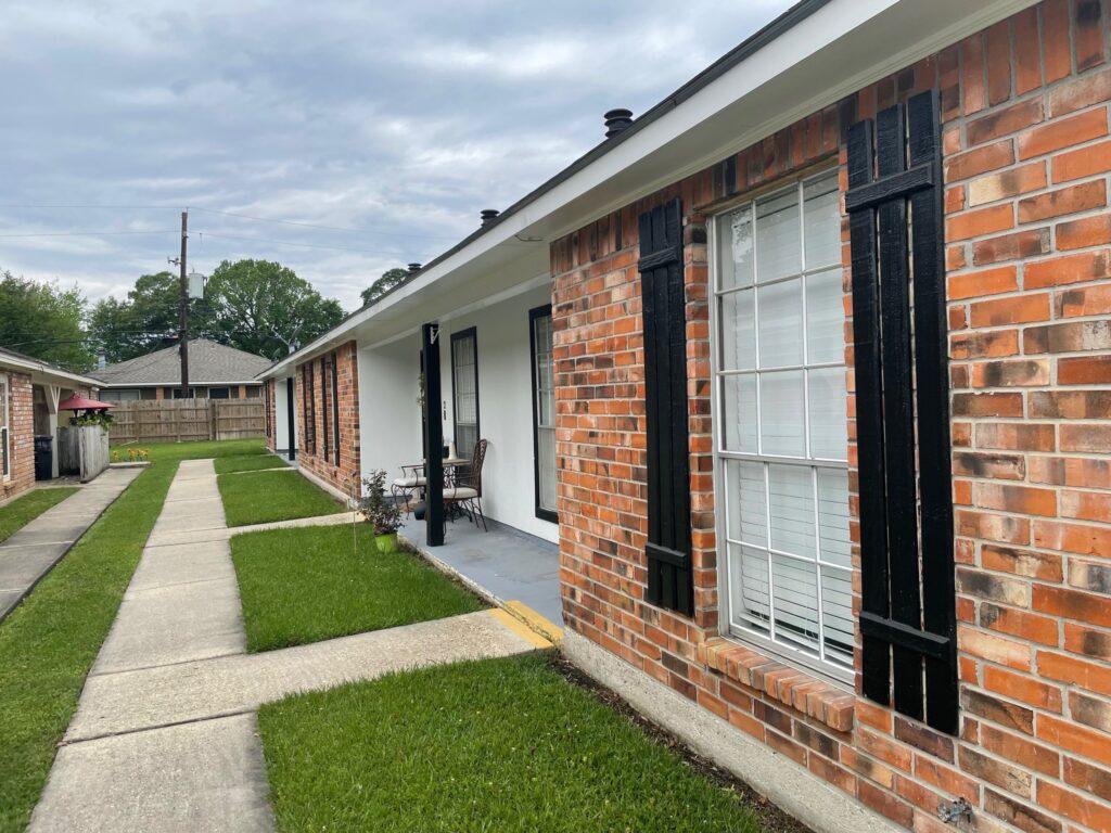 A brick house with black shutters and a concrete walkway in front, bordered by a neatly trimmed lawn. A wooden fence is visible in the background, showcasing prime Baton Rouge real estate by Lewis Companies, offering excellent property for lease.