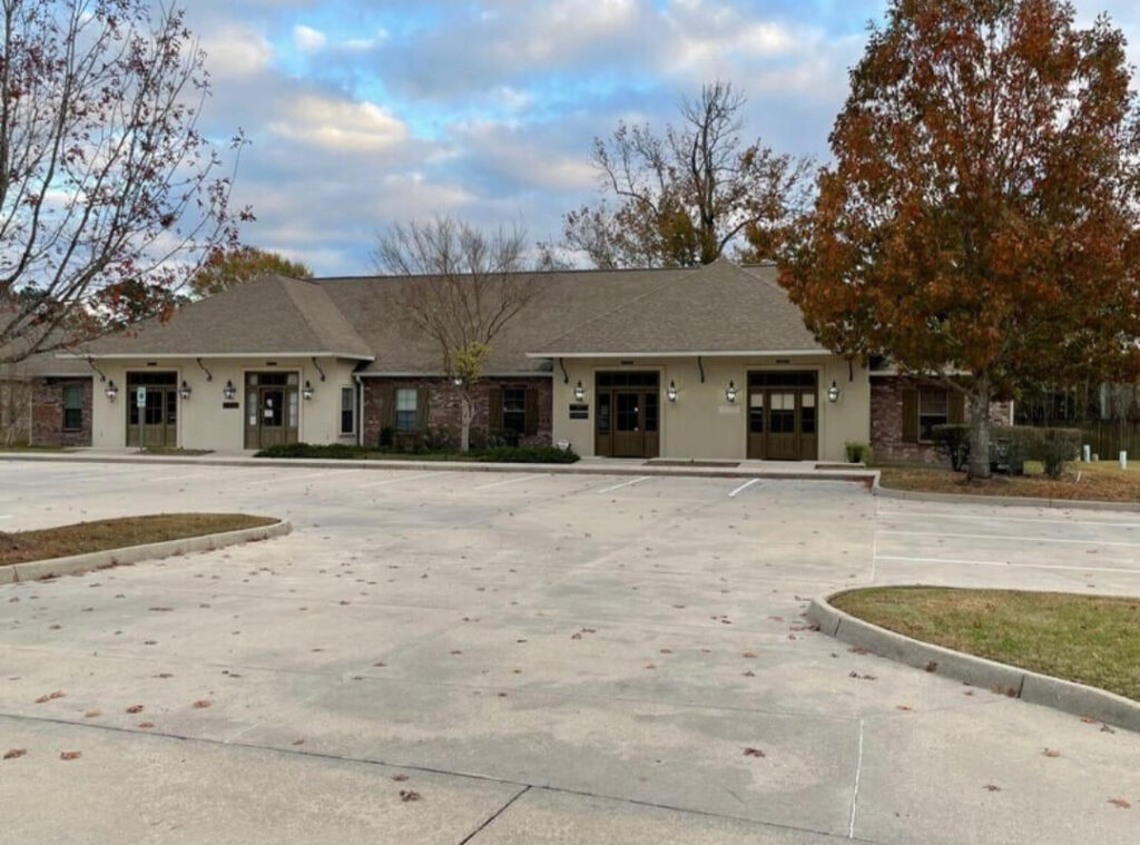 A building with a light brown exterior and multiple entrances, managed by Lewis Companies. It is surrounded by autumn-colored trees and an empty parking lot in the foreground, offering a prime property for lease.