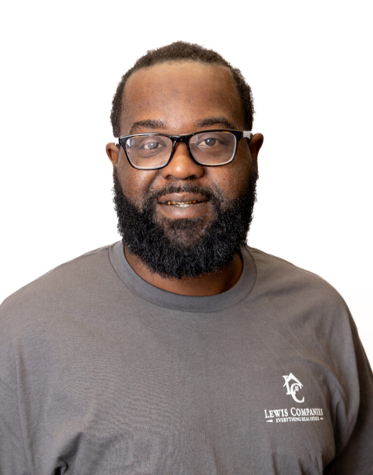 A man with a beard wearing glasses and a gray Lewis Companies shirt smiles at the camera against a white background, reflecting the professional touch of Baton Rouge real estate.