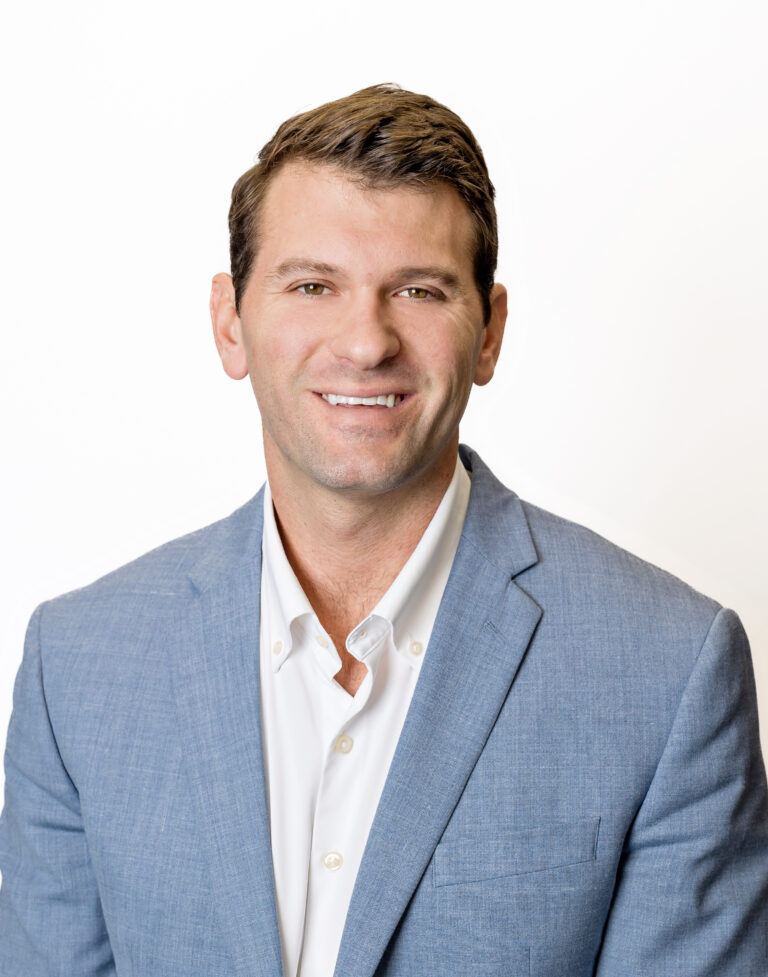 A man in a light blue blazer and white shirt is smiling, set against a white background. As a representative of Baton Rouge real estate, his friendly demeanor is perfect for showcasing properties for lease.