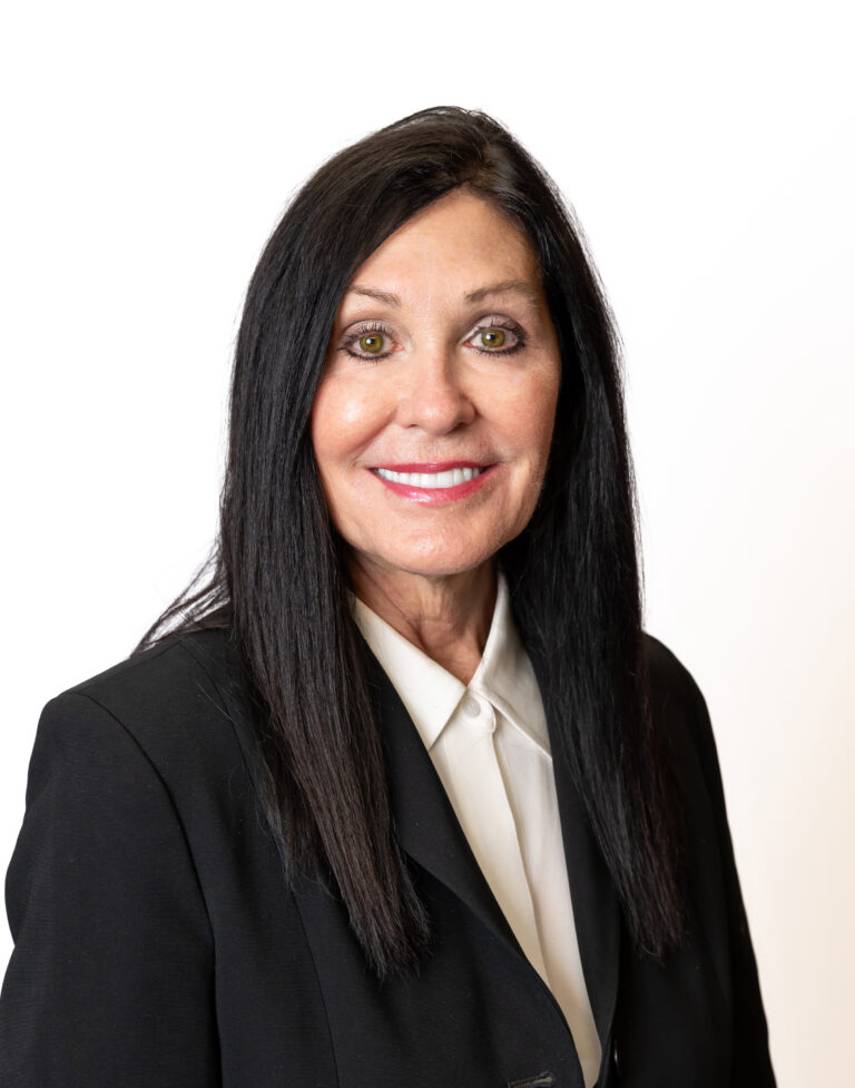 A woman with long black hair, wearing a black blazer and white shirt, is smiling against a plain white background, representing Baton Rouge real estate for the Lewis Companies.