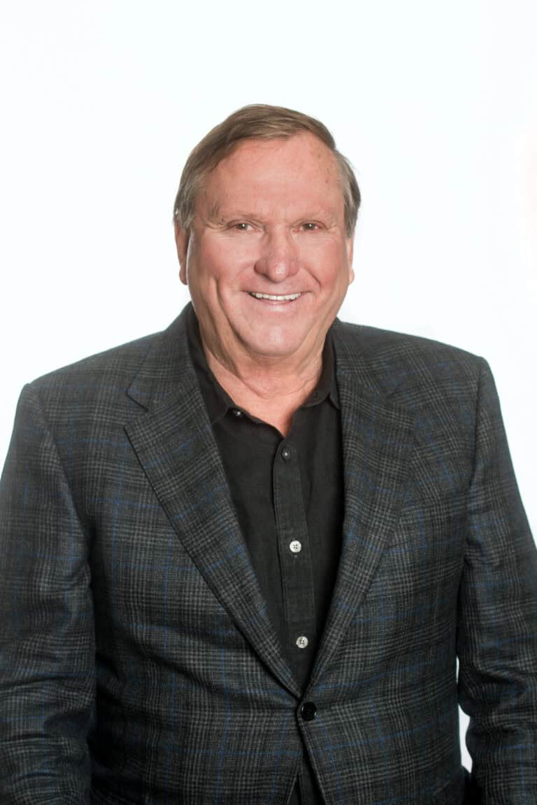 A man in a dark checkered blazer and black shirt stands against a plain background, smiling at the camera.
