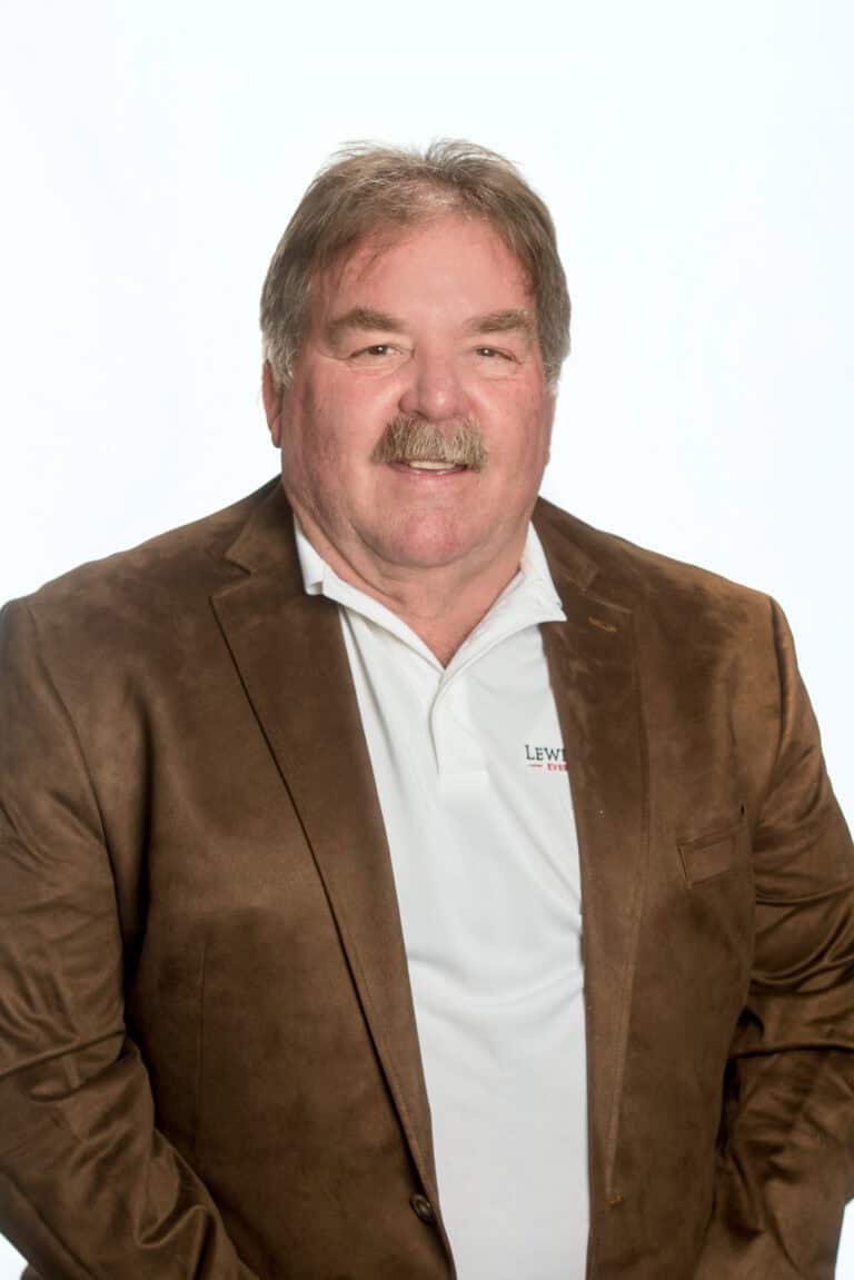 A man with gray hair and a mustache is wearing a brown blazer over a white polo shirt, standing against a plain white background.