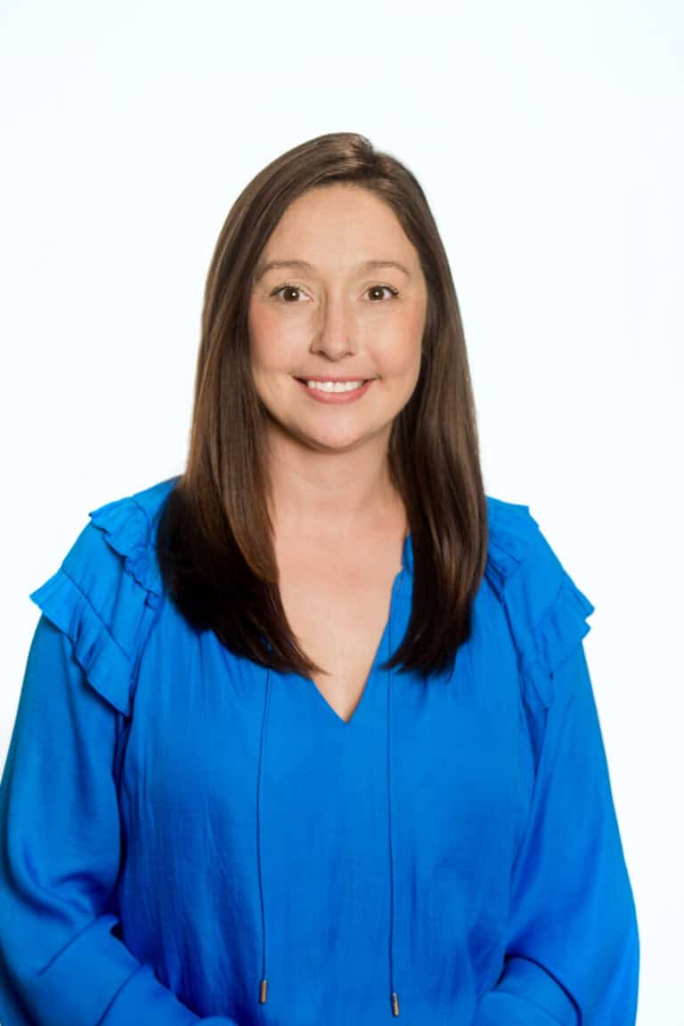 A woman with straight brown hair wearing a blue blouse smiles at the camera against a plain white background.